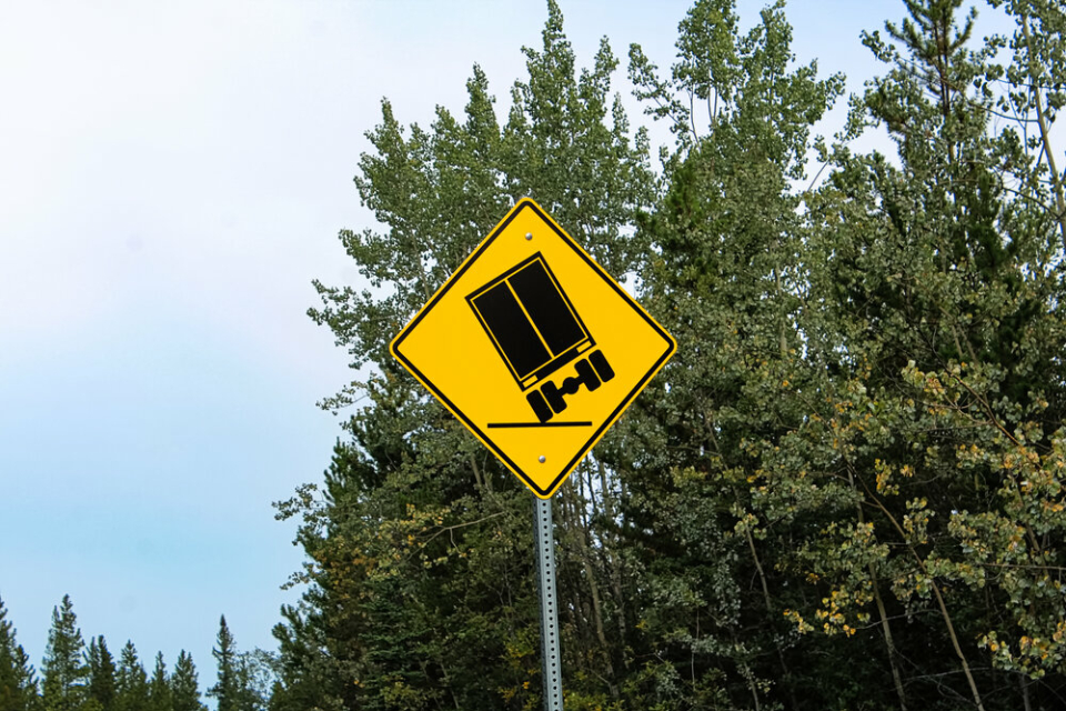 A yellow warning road sign with a symbol of an overturning truck on it, and green trees in the background.
