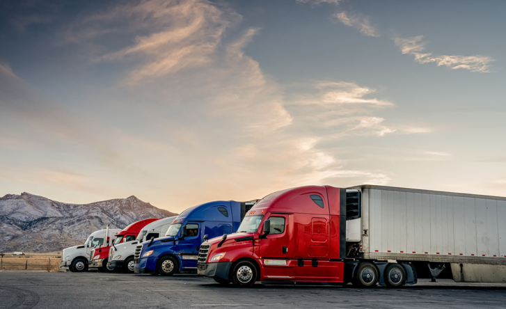 Five trucks are parked and lined up for the night, their cabins are red, blue or white. There are mountains in the background and it's twilight.