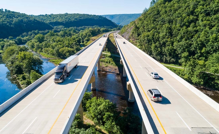 A photo of two flyover highways which are built across a river with forest on either side. There's a truck on the left and two cars on the right, as well as some cars in the background.
