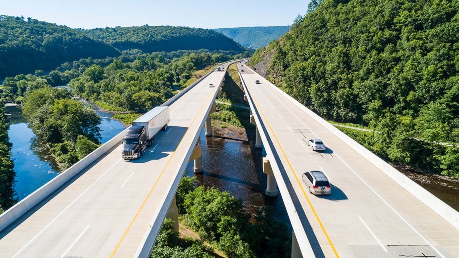 A photo of two flyover highways which are built across a river with forest on either side. There's a truck on the left and two cars on the right, as well as some cars in the background.