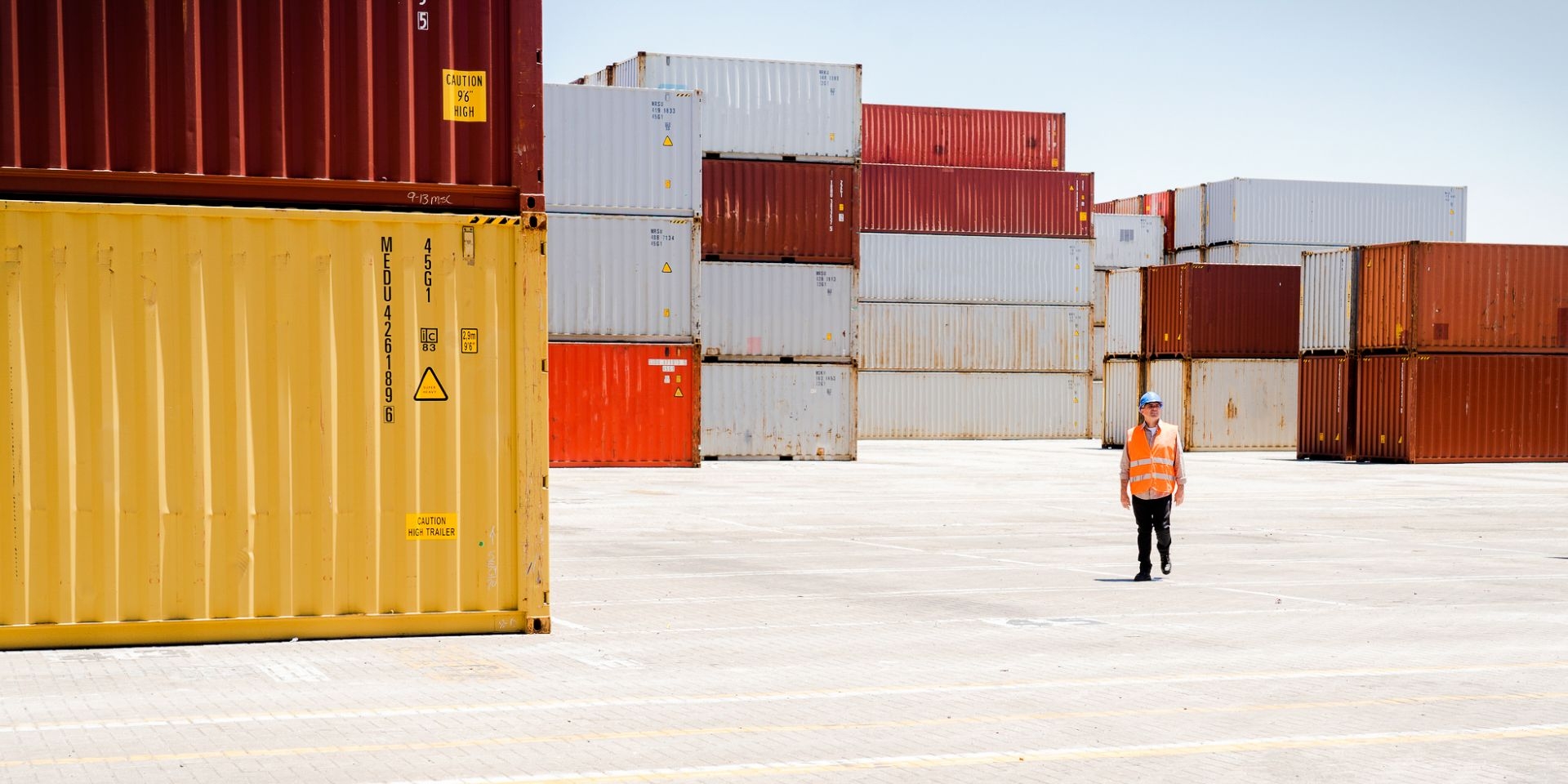A photo of a man in orange high vis and a hard hat, walking through a ship yard full of orange, red and white containers.