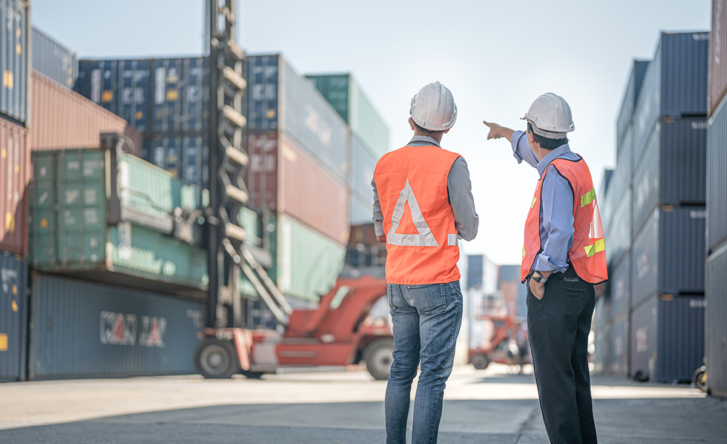 Two men in hard hats and high vis stand surrounded by shipping containers, one is pointing into the distance while the other looks on.