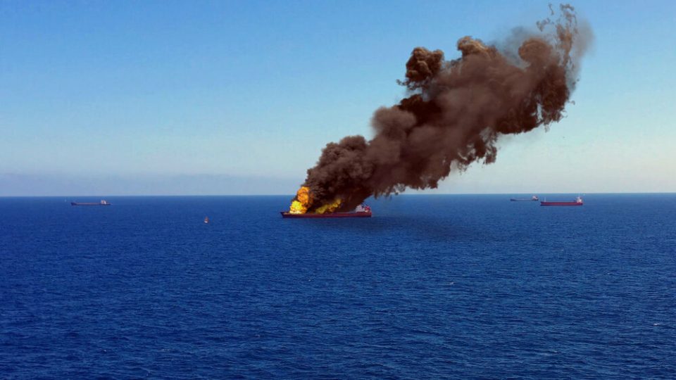 A landscape of the sea with a marine cargo boat in the middle, which is on fire. Large yellow flames send black clouds of smoke up into the air.