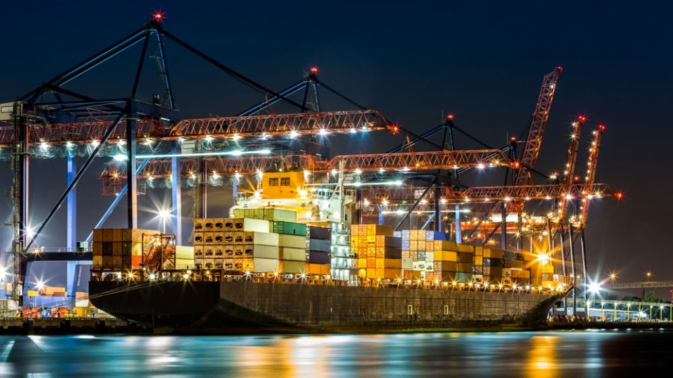 A photo of a marine cargo ship lit up at night, while it docks. There are several cranes operating and the ship yard is lit up.