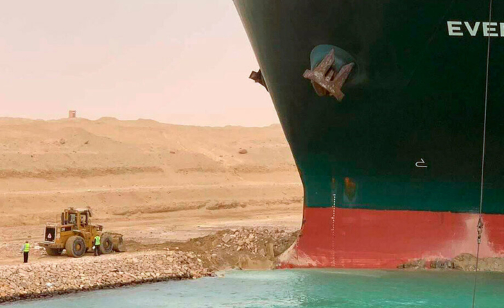 A close-up shot of the marine cargo vessel the Ever Given, stuck in the side of the Suez Canal. A digger is parked next to it.