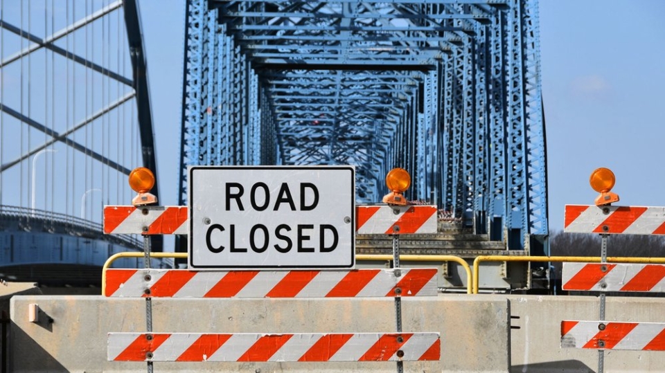 A photo of a bridge with a large blockade across it and a sign that says 'road closed'.