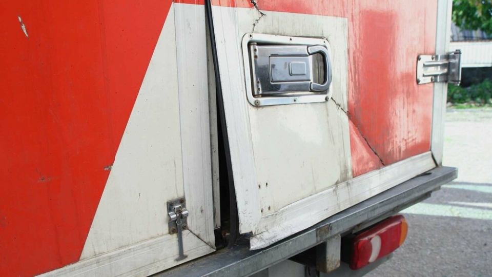 A photo showing the corner of a red and white cargo truck where it's clear that entry has been forced - the door is bent and a crack runs through the cover.