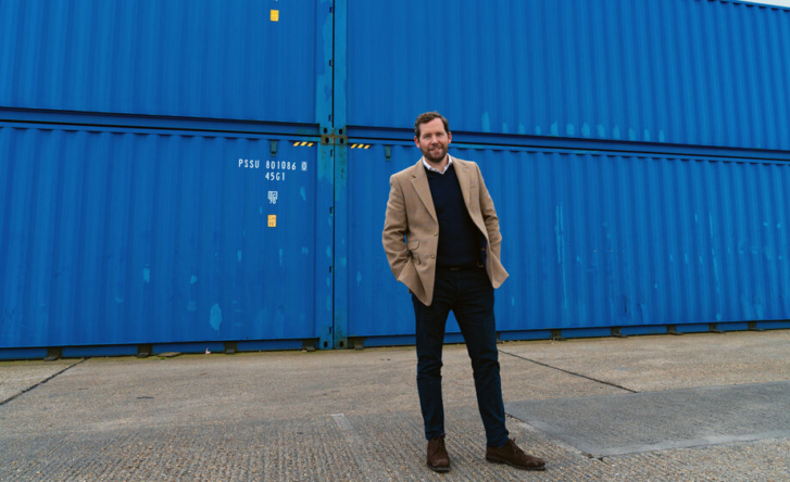 A photo of Johnny McCord in a light brown jacket, black jumper and white shirt, standing with his hands in his pockets in front of several blue shipping containers.