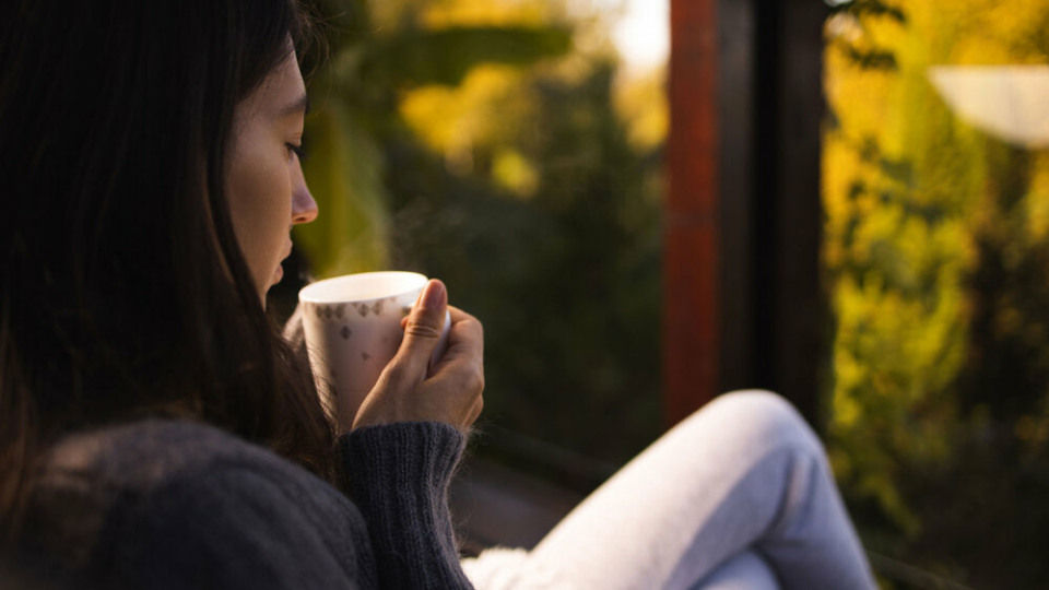 A close up of a woman holding a hot drink in a mug, with her legs crossed as she looks out of the window.