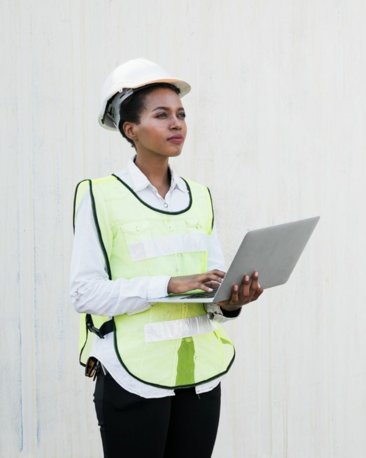 A woman in high vis and a hard hat is standing in front of a white wall with a laptop, looking into the distance.