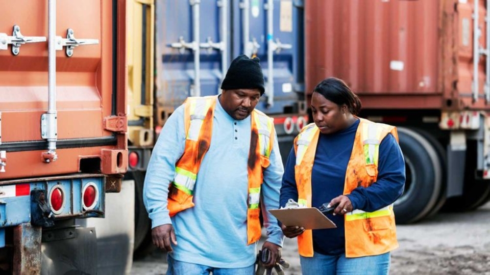 A photo of two people in a shipyard looking at a clipboard, both wearing orange high vis.