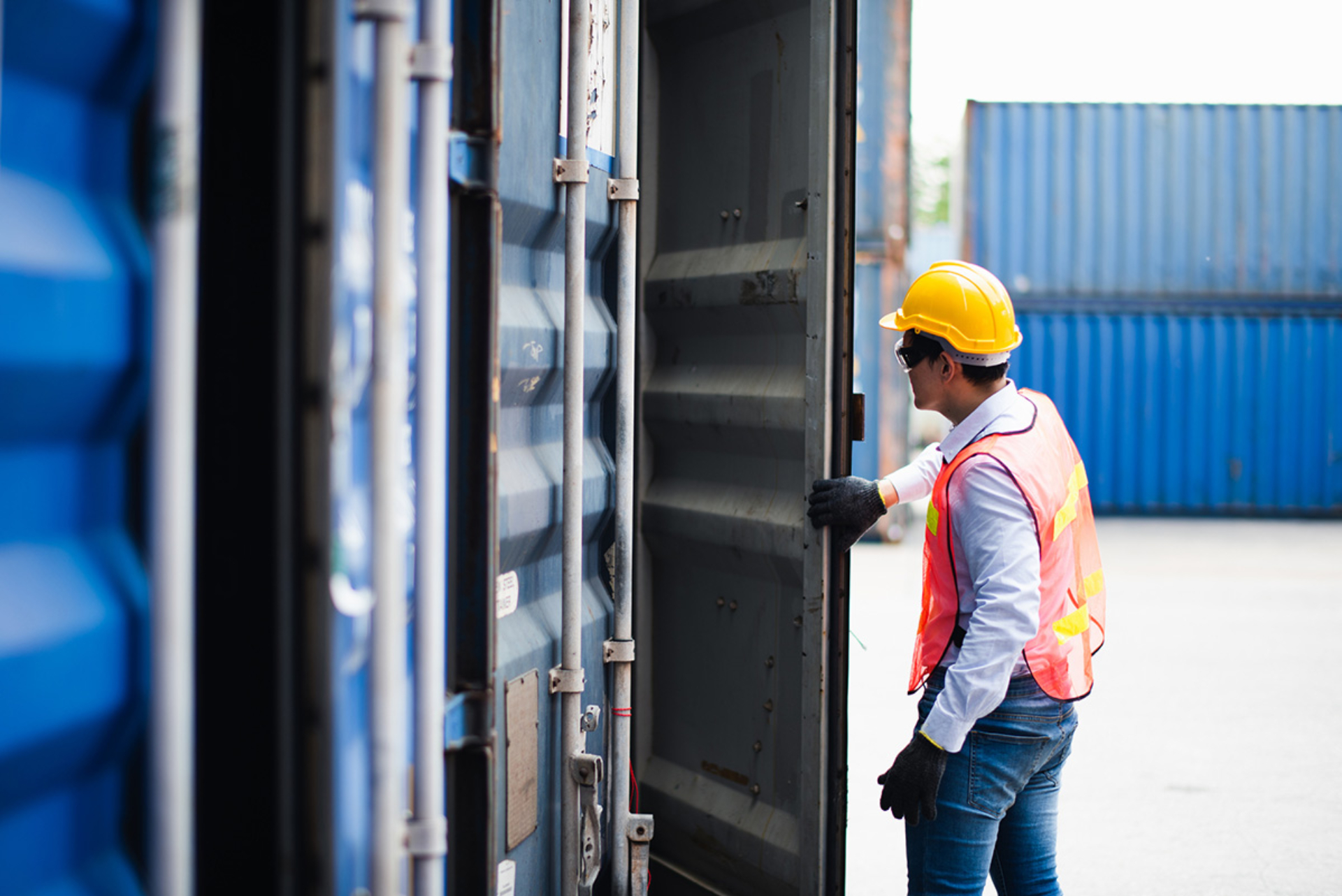 A man in protective gear is opening a shipping container door in a ship yard. There are blue shipping containers in the background. =
