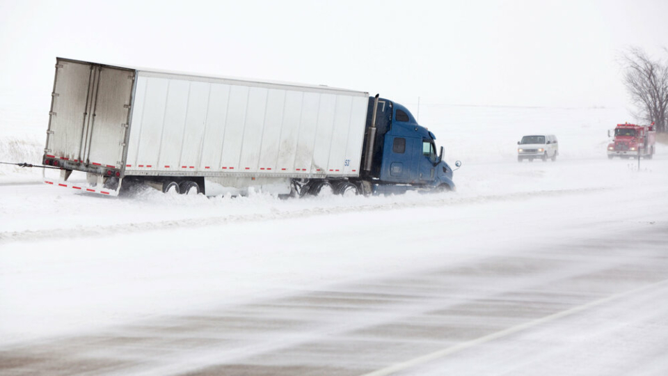 A cargo truck that has come off the road in the snow, with a blue cabin. Two cars are approaching it from the other direction.