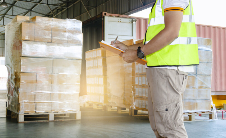 A shot from the shoulders down of a man standing in a warehouse full of wrapped pallets. He is wearing high vis and holding an orange clipboard.
