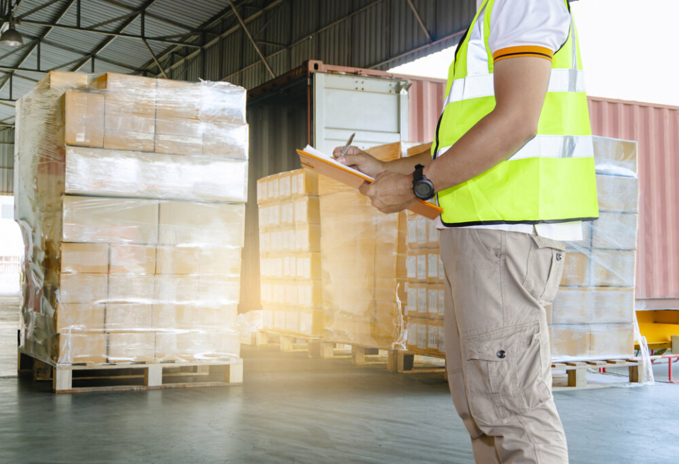 A shot from the shoulders down of a man standing in a warehouse full of wrapped pallets. He is wearing high vis and holding an orange clipboard.