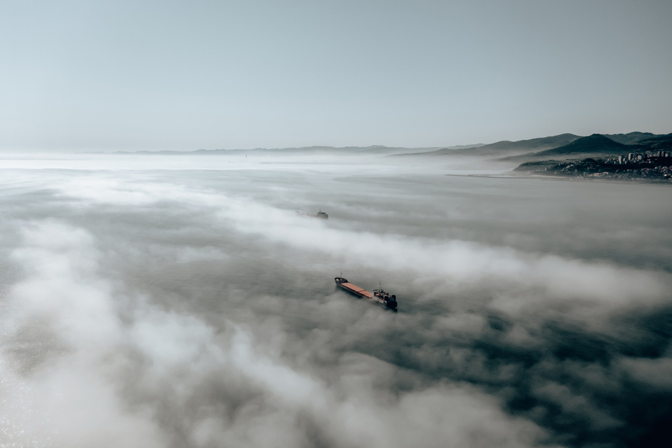 A landscape photo of the sea, which is almost completely covered by fog. You can see the coastline on the right and one cargo boat emerging through the mist.