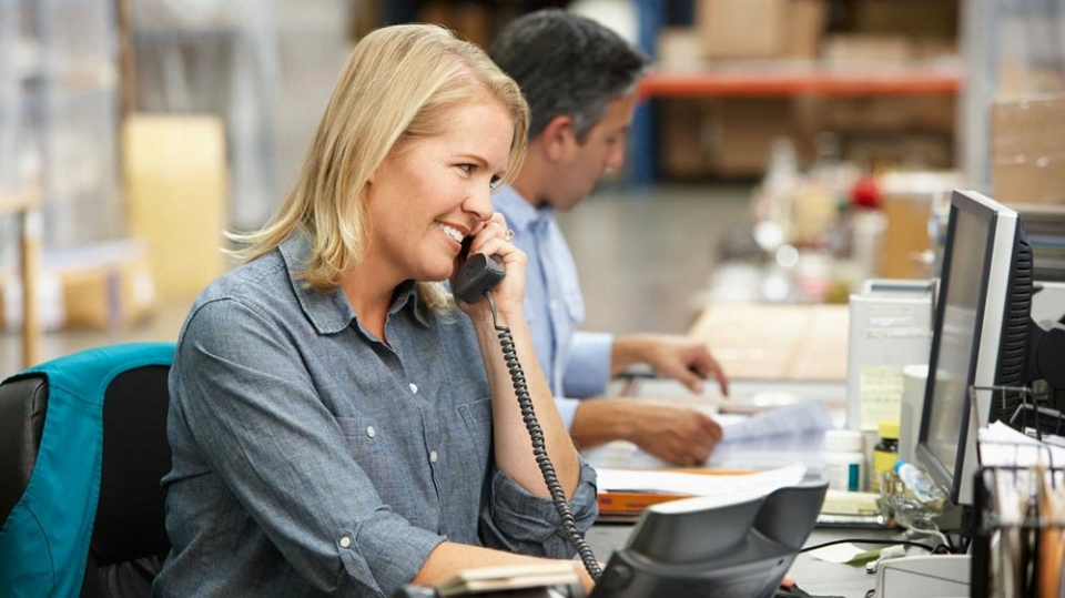 A photo of a blonde woman at a desk on the phone, in a grey shirt. A man is looking at some paperwork behind her.