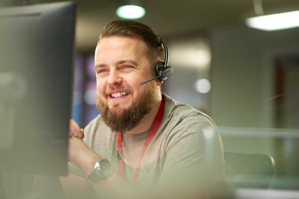 A man wearing a headset sitting at a desk and smiling, he's wearing a red lanyard and a grey tshirt,