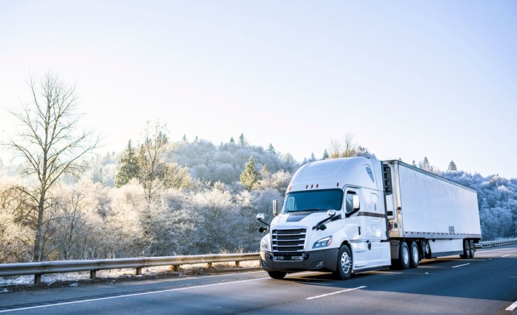 A white cargo truck driving along a highway on a sunny day, with winter scenery alongside it on the left.
