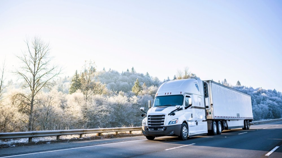 A white cargo truck driving along a highway on a sunny day, with winter scenery alongside it on the left.