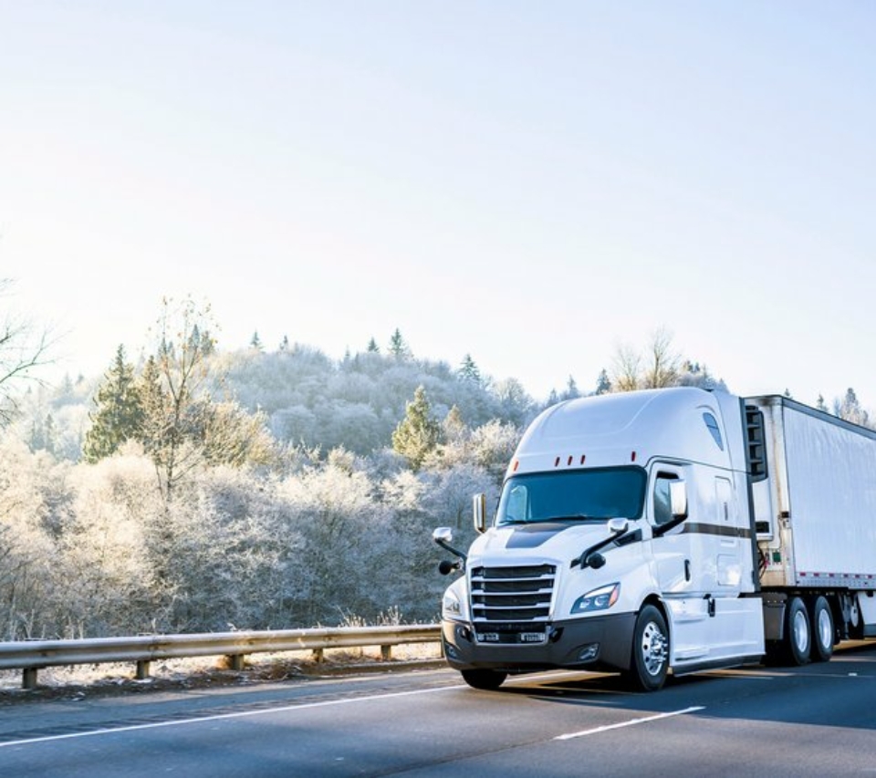 A white cargo truck driving along a highway on a sunny day, with winter scenery alongside it on the left.