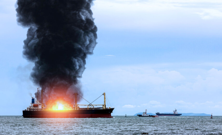 A photograph of a marine cargo ship at sea, which is on fire. Large plumes of black smoke are coming off it, and several boats can be seen in the background.