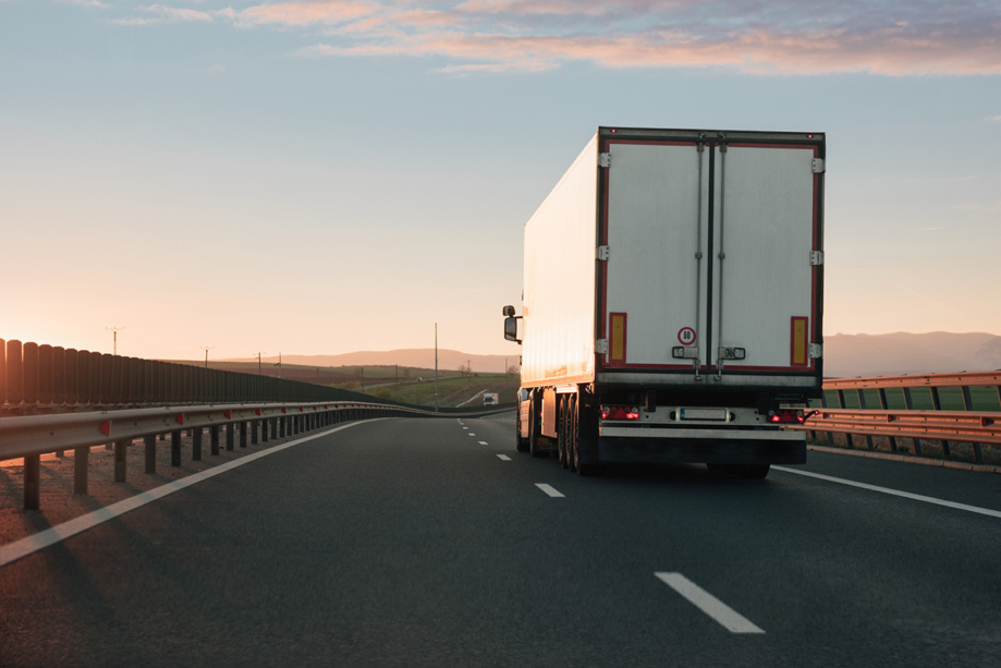 A white cargo truck is pictured from behind as it drives along the right lane of a highway. The sun is setting on the left.