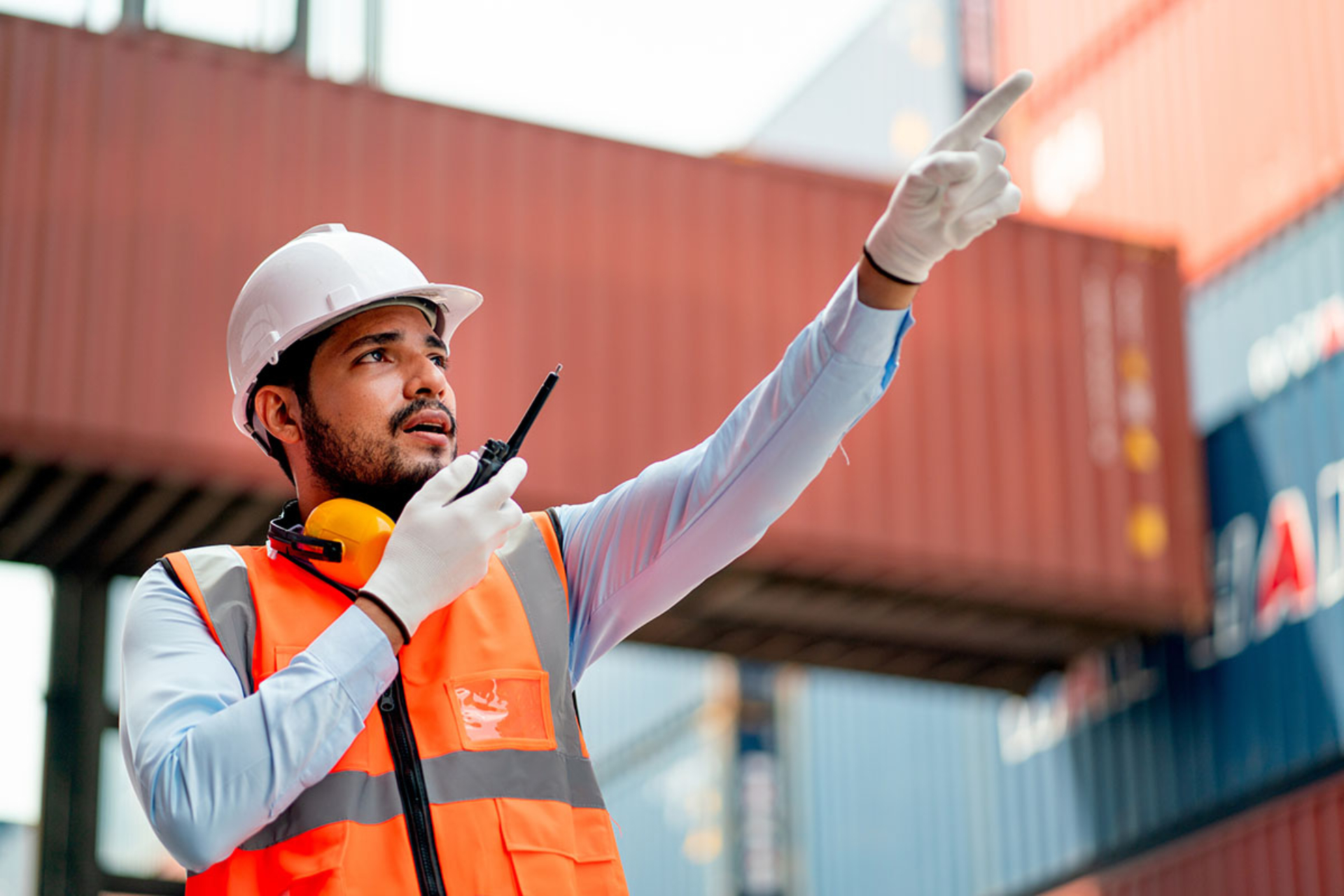A man in protective gear stands in a ship yard. He is talking to someone over a radio and pointing at something in the distance.