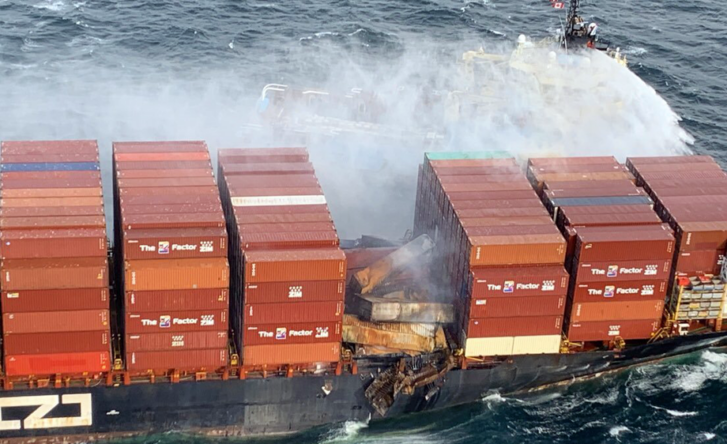 A photo of a marine cargo ship covered in shipping containers, some of which are damaged by water or fire. A boat nearby is trying to help.