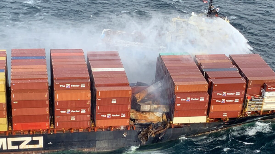 A photo of a marine cargo ship covered in shipping containers, some of which are damaged by water or fire. A boat nearby is trying to help.