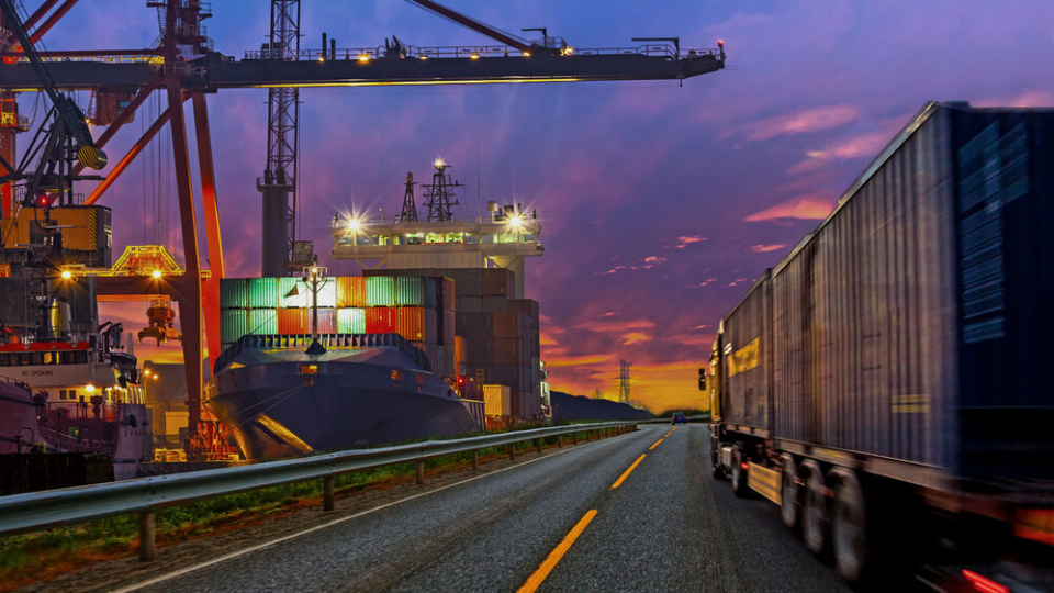 A black cargo truck driving down a highway at sunset, past a lit up ship yard.