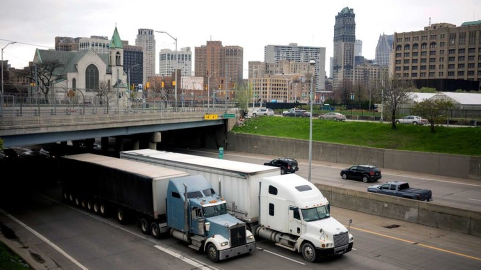 An image of a highway in a city with two trucks in the foreground, one blue on white.