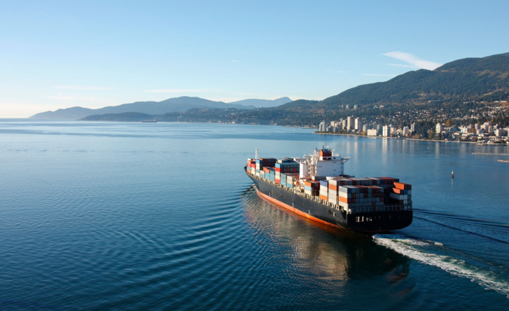 A heavily loaded cargo ship is at sea. On the right you can see a coastline of a city, with green hills and mountains behind it.