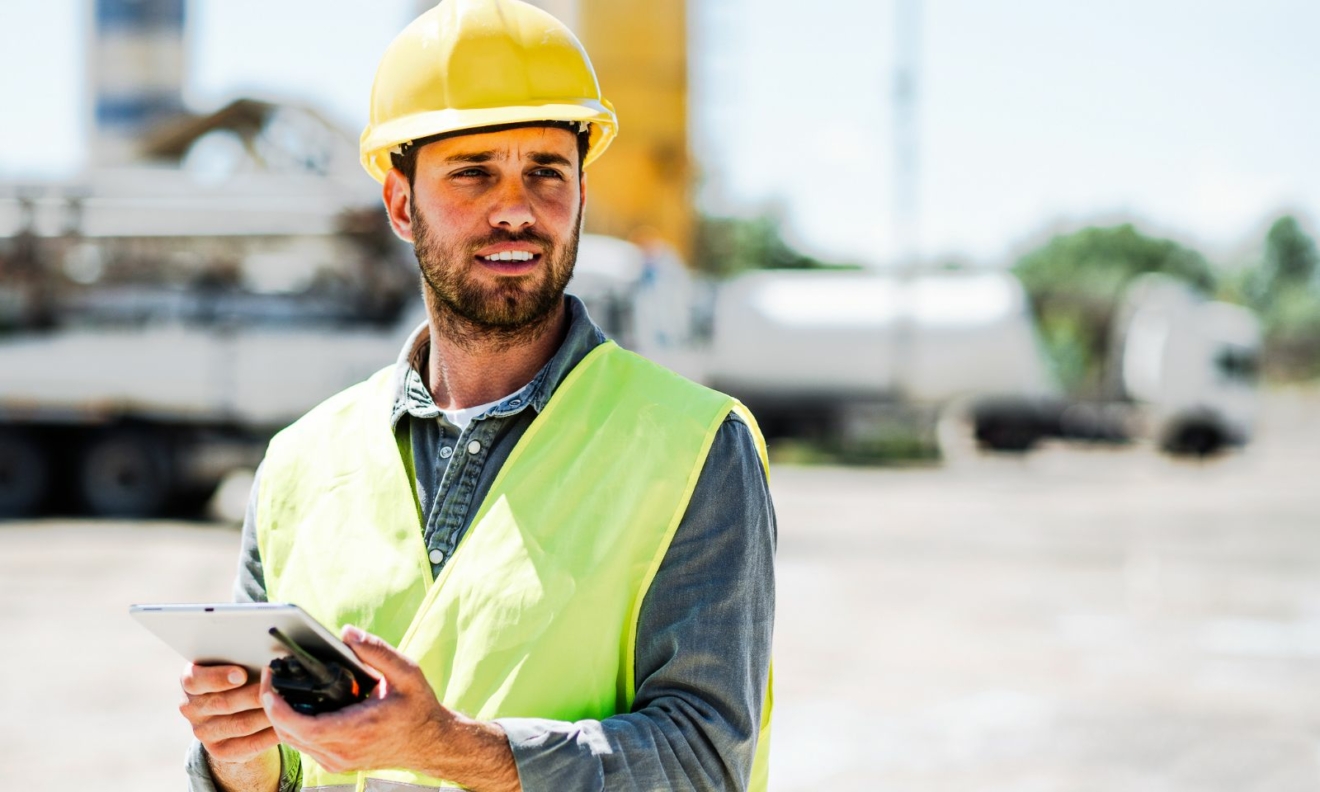 A photo of a man in a yellow hard hat and high vis, holding a tablet and a handheld radio.