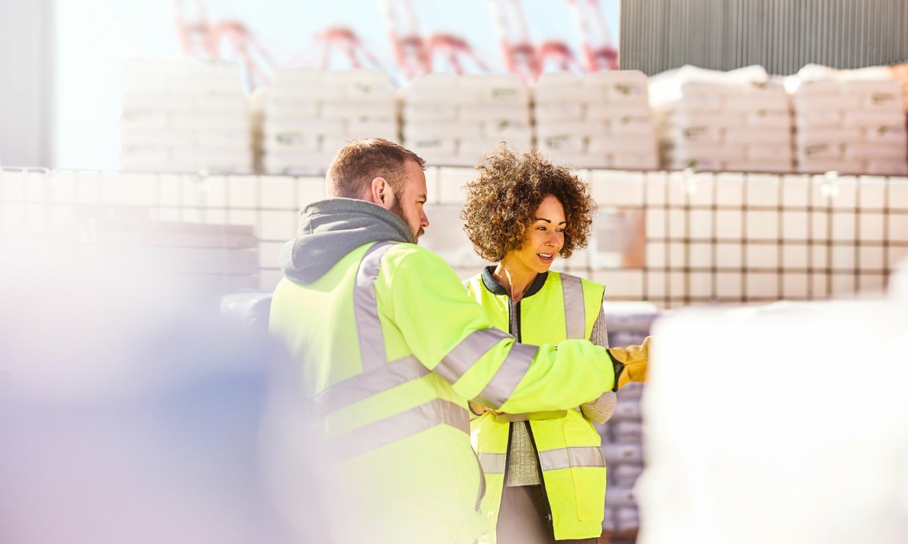 An image of a man and a woman in high vis jackets, surrounded by pallets full of stick. They are looking at something out of sight.